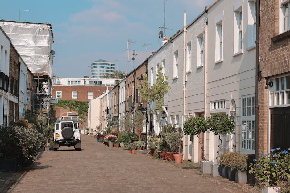 Image depicting a narrow residential street in Ealing with a white van parked on the cobblestone road, which is lined with various potted plants and small trees outside terraced houses. The houses have brick and white facades, with some windows featuring decorative leaded glass and small porches. Adjacent to the street, a delivery trolley with wrapped furniture or boxes is visible, indicating a home relocation or moving process. The scene is captured during daylight hours with a clear sky, and in the background, there are modern office buildings visible behind the residential area. This setting illustrates the logistical considerations faced by Ealing Removals when conducting furniture transport and packing during a house move in a tight street environment, highlighting the importance of careful planning and maneuvering equipment such as trolleys and vans during the loading process.
