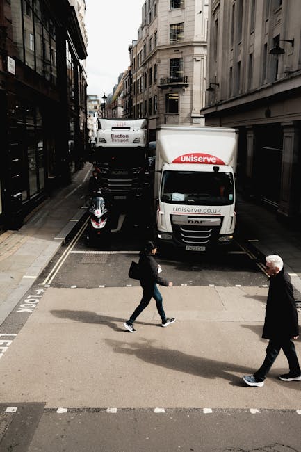 A narrow urban street scene depicting a home relocation process during furniture transport and packing and moving activities. Two large white box trucks with 'uniserve.co.uk' branding are parked along the street; the first truck is positioned directly in front of a building entrance, while a second larger truck is parked behind it on the same road. An electric scooter is parked on the pavement beside the trucks. In the foreground, two individuals are crossing the street on a zebra crossing—one person dressed in dark clothing and carrying a bag, and an elderly man in a dark coat and white hair. The street is lined with multi-storey buildings with classic architecture, some with balconies and fire escapes, illuminated by natural daylight. The scene captures the logistical aspects of moving services in an urban environment, supported by Ealing Removals' expertise in handling tight streets and complex loading areas.