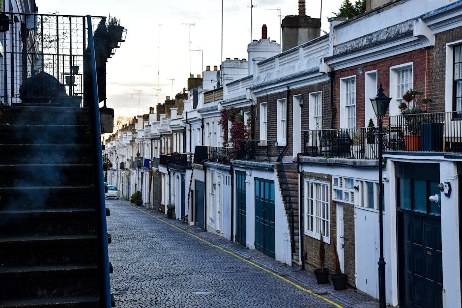 Image depicting a narrow residential street in Ealing with a white van parked on the cobblestone road, which is lined with various potted plants and small trees outside terraced houses. The houses have brick and white facades, with some windows featuring decorative leaded glass and small porches. Adjacent to the street, a delivery trolley with wrapped furniture or boxes is visible, indicating a home relocation or moving process. The scene is captured during daylight hours with a clear sky, and in the background, there are modern office buildings visible behind the residential area. This setting illustrates the logistical considerations faced by Ealing Removals when conducting furniture transport and packing during a house move in a tight street environment, highlighting the importance of careful planning and maneuvering equipment such as trolleys and vans during the loading process.