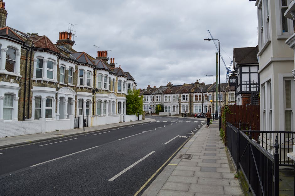 A quiet residential street with Victorian-style terraced houses featuring bay windows and chimneys, situated on Pitshanger Lane in Ealing. The pavement on the right side is made of concrete slabs, with a black metal fence running alongside the front of some houses. Overcast skies cast diffuse lighting over the scene, and a single pedestrian is walking away from the camera. No moving vehicles or loading activity are visible in this image, which captures a calm moment prior to home relocation or moving activities. Ealing Removals, a professional removals company, operates in this area and may utilize streets like this for furniture transport, packing and moving, or access planning related to house removals and parking arrangements.